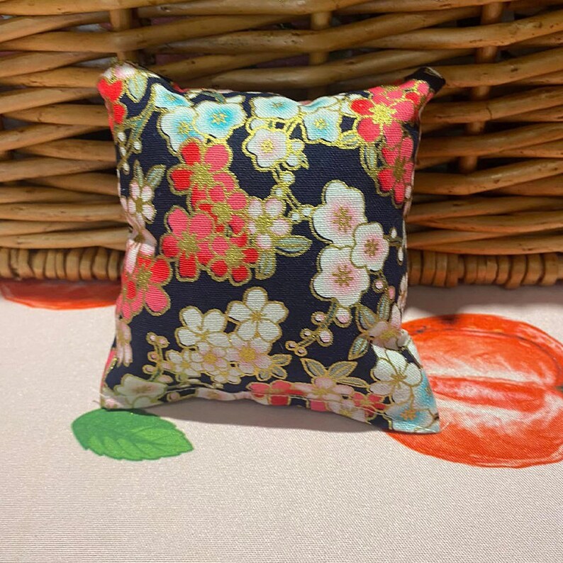 May include: A small, square cushion with a navy blue background and a floral pattern in shades of pink, red, and gold. The cushion is made of fabric and has a soft, plush texture.