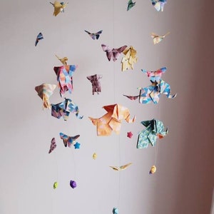 May include: A handmade baby mobile featuring origami elephants and butterflies in various colors and patterns. The mobile hangs from a wooden hoop and includes small star and geometric shapes, creating a whimsical nursery decoration.