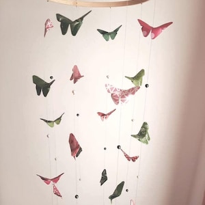 May include: A wooden ring mobile with green and pink butterflies hanging from it. The butterflies are made of paper and have a floral pattern. The mobile is decorated with black and white beads.