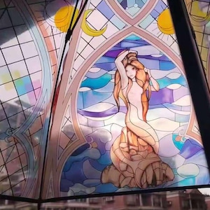 Puede incluir: Un paraguas abierto con un diseño de vidriera que presenta una figura de sirena. La obra de arte utiliza tonos de naranja, azul y morado. El marco del paraguas es negro, y el fondo muestra edificios y árboles.