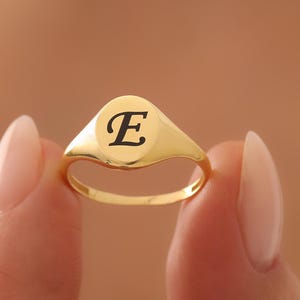 May include: A gold signet ring with a polished finish. The ring features a rounded, oval face with the letter "E" in black script. The ring is held up by a person's fingers.