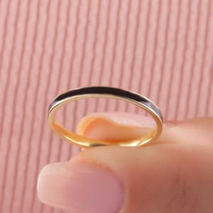 Könnte beinhalten: Ein goldener Ring mit einem dünnen schwarzen Emailleband. Der Ring wird von einem Finger vor einem rosa Hintergrund gehalten. Der Ring ist ein schlichtes, elegantes Design.