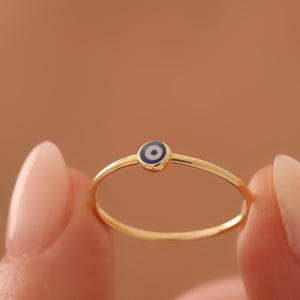 Könnte beinhalten: Ein dünner Goldring mit einem kleinen, kreisförmigen Nazar-Amulett in der Mitte. Das Amulett besteht aus einer dunkelblauen Pupille, die von einem weißen Ring und einem dünnen blauen Rand umgeben ist, auf dem polierten gelben Goldring.