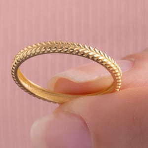 May include: A thin gold band ring featuring a continuous, repeating etched leaf or wheat pattern along the entire outer surface. The metal has a polished, reflective finish, and the design consists of small, angled, V-shaped grooves.