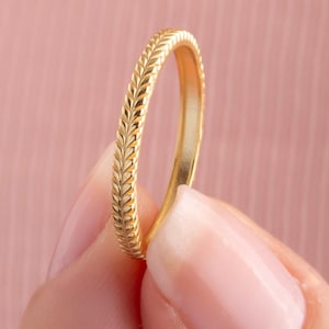 Könnte beinhalten: Ein goldfarbener Ring mit einem detaillierten Weizen- oder Blättermuster um das Band. Der Ring wird vor einem weichen rosa Hintergrund gehalten, der sein aufwendiges Design und seinen metallischen Glanz hervorhebt. Der Ring ist ein Schmuckstück.