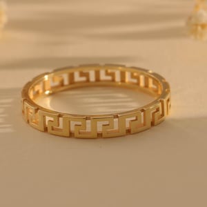 Op de afbeelding: Een goudkleurige metalen ring met een doorlopend uitgesneden Grieks sleutelpatroon rondom de gehele band. De ring heeft een gepolijste afwerking en een uniforme breedte, met een geometrisch ontwerp dat zich consistent herhaalt.