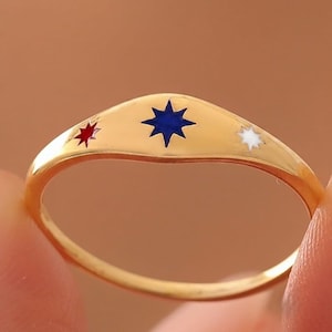 Op de afbeelding: Een goudkleurige ring met een gladde, gebogen band en drie ingelegde stervormen aan de bovenzijde. De middelste ster is donkerblauw, geflankeerd door een kleinere rode ster aan de linkerkant en een kleinere witte ster aan de rechterkant.