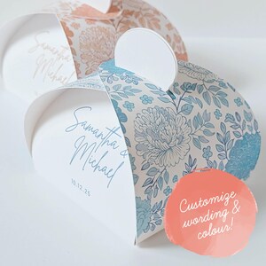 May include: Three heart-shaped favour boxes. Two boxes feature floral patterns in blue and peach. The third box is white with the names "Samantha & Michael" and the date "10.12.25" printed in script. A peach circle says "Customize wording & colour!"