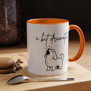 May include: White ceramic mug with an orange interior and handle. The mug has a black illustration of a dog and the phrase "a bit dramatic" in black script. A spoon and a macaron are on a wooden surface.