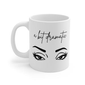 Può includere: Tazza in ceramica bianca con un'illustrazione nera di occhi stilizzati e la frase "a bit dramatic" in corsivo nero. La tazza ha un manico curvo e una finitura lucida.