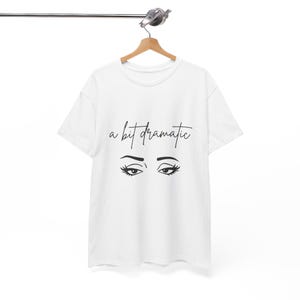 Può includere: T-shirt bianca con la scritta "a bit dramatic" in corsivo nero sopra un paio di ciglia e sopracciglia nere stilizzate. La t-shirt è appesa a una gruccia di legno.