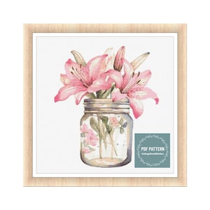 Peut inclure: Une peinture à l'aquarelle de lys roses dans un bocal en verre transparent avec un couvercle brun rustique. Le bocal est posé sur un fond blanc. La peinture est encadrée dans un cadre en bois clair. Le texte "PDF PATTERN CottageCoreStitches" est dans le coin inférieur droit de l'image.