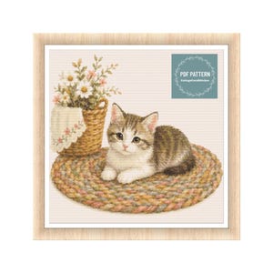 Op de afbeelding: Een kruissteekpatroon met een bruin-witte cyperse kitten die op een rond gevlochten kleed ligt. Naast de kitten staat een gevlochten mand gevuld met witte en roze bloemen en een witte doek. De tekst PDF PATTERN CottageCoreStitches is zichtbaar.