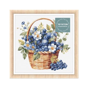 Può includere: Un dipinto ad acquerello di un cesto pieno di mirtilli e fiori bianchi. Il cesto è marrone e intrecciato. I mirtilli sono blu e i fiori sono bianchi con centri gialli. Il dipinto è incorniciato in una cornice di legno chiaro. Il testo "PDF PATTERN CottageCoreStitches" è nell'angolo in alto a destra dell'immagine.