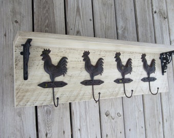 May include: White wooden shelf with four black metal rooster hooks. The shelf has a distressed white finish and is mounted on a wood wall.