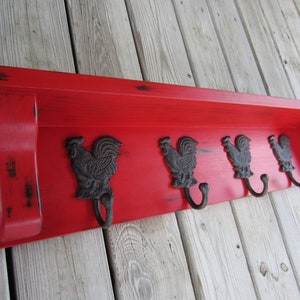 May include: Red wooden shelf with four rooster-shaped hooks. The shelf has a distressed finish and is mounted on a wood wall.