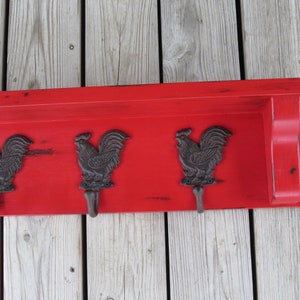 May include: Red wooden shelf with three rooster-shaped hooks. The shelf has a distressed finish and is mounted on a wood wall.