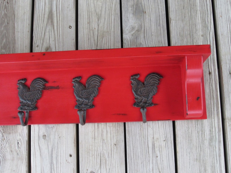 May include: Red wooden shelf with three rooster-shaped hooks. The shelf has a distressed finish and is mounted on a wood wall.
