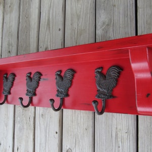 May include: Red wooden shelf with four black rooster hooks. The shelf has a distressed finish and is mounted on a wood wall.