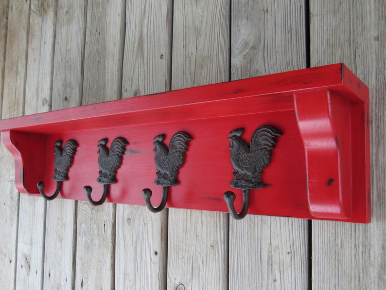May include: Red wooden shelf with four black rooster hooks. The shelf has a distressed finish and is mounted on a wood wall.