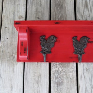 May include: Red wooden shelf with three black rooster hooks. The shelf has a distressed finish and is mounted on a wood wall.