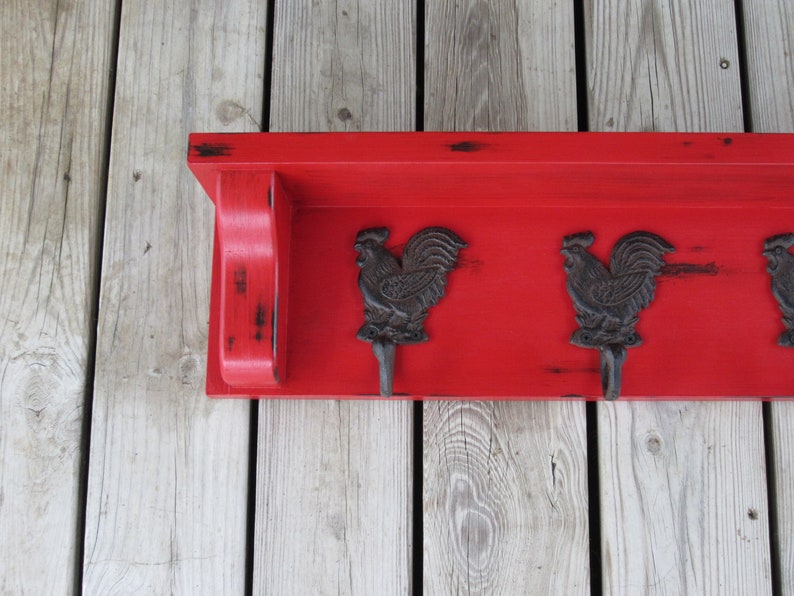 May include: Red wooden shelf with three black rooster hooks. The shelf has a distressed finish and is mounted on a wood wall.