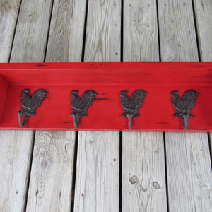 May include: Red wooden shelf with four black rooster hooks. The shelf has a distressed finish and is mounted on a wood wall.