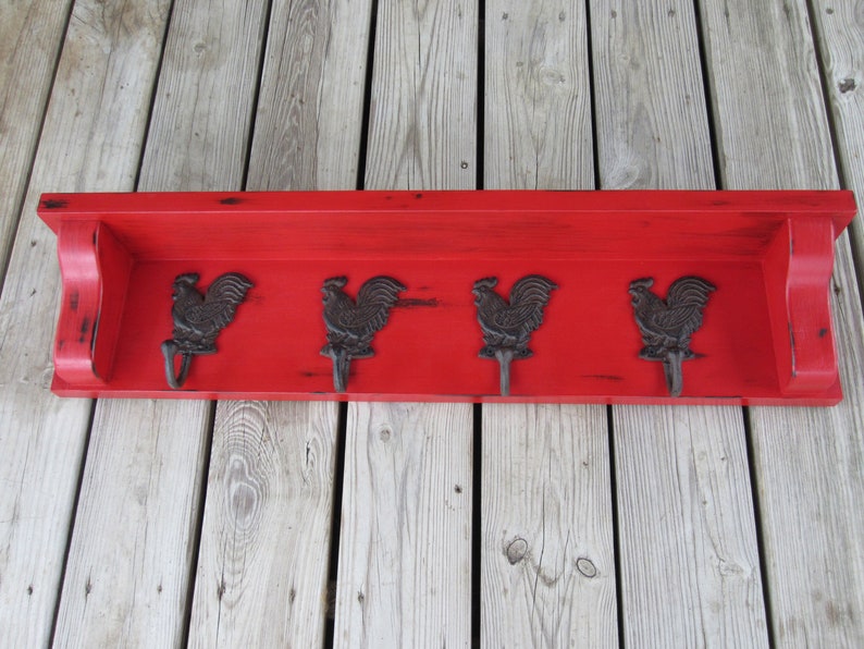 May include: Red wooden shelf with four black rooster hooks. The shelf has a distressed finish and is mounted on a wood wall.