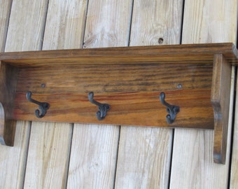 May include: A wooden shelf with three black metal hooks. The shelf is stained a dark brown color and has a rustic look.