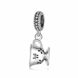 Könnte beinhalten: Silberner Anhänger in Form einer Teetasse mit floralem Design. Der Anhänger hat eine perlenartige Schlaufe zum Anbringen an einem Armband oder einer Halskette.