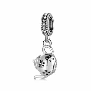 Könnte beinhalten: Ein silberner Charm mit einem Teekannen-Design und floralen Aussparungen. Der Charm hat eine dekorative Oberseite mit kleinen, herzförmigen Details. Die Teekanne hat einen Ausguss und einen Griff und ist an der dekorativen Oberseite aufgehängt.