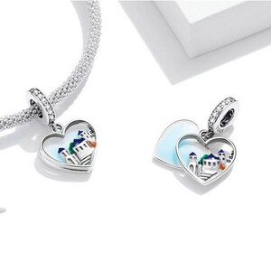 May include: Two silver charm pendants in the shape of hearts. The charms feature a white enamel background with a colorful depiction of a small town inside. One charm is a single heart, while the other is two hearts that open to reveal the town.