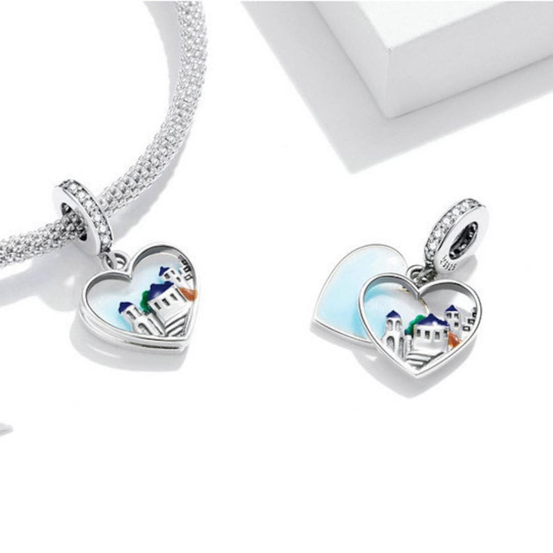 May include: Two silver charm pendants in the shape of hearts. The charms feature a white enamel background with a colorful depiction of a small town inside. One charm is a single heart, while the other is two hearts that open to reveal the town.