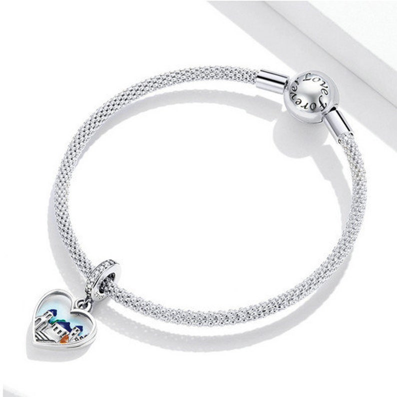 May include: A silver charm bracelet with a heart-shaped charm featuring a colorful cityscape. The bracelet has a mesh design and a clasp that reads "Love Forever".