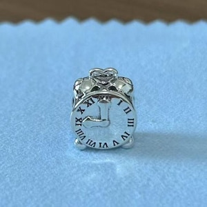 May include: Silver charm in the shape of an alarm clock with Roman numeral markings. The clock face has a silver hand pointing to the number 12.