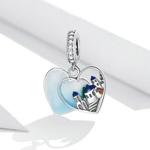 May include: A silver charm in the shape of two interlocking hearts. The hearts are decorated with a white enamel background and a colorful depiction of a small town with buildings. The charm is adorned with clear cubic zirconia stones.