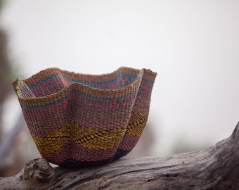 May include: A woven basket with a colorful geometric pattern in shades of pink, blue, green, and yellow. The basket is sitting on a piece of driftwood.
