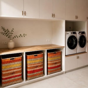 May include: A laundry room featuring white cabinetry with brass handles, a wooden countertop, and three rectangular woven baskets with horizontal stripes in red, orange, and yellow. A white vase with green branches sits on the counter.