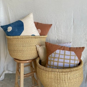 Puede incluir: Dos cestas tejidas con asas, una encima de la otra, llenas de almohadas decorativas de colores. La cesta superior est&aacute; llena de una almohada blanca con dise&ntilde;os abstractos azules y rosas, una almohada color &oacute;xido y una almohada blanca con un dise&ntilde;o abstracto azul. La cesta inferior est&aacute; llena de una almohada color &oacute;xido con un dise&ntilde;o geom&eacute;trico morado claro y marr&oacute;n y una almohada blanca.