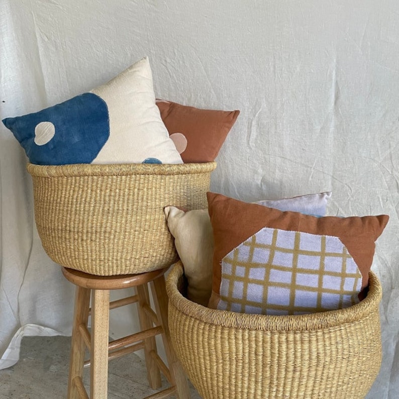 Puede incluir: Dos cestas tejidas con asas, una encima de la otra, llenas de almohadas decorativas de colores. La cesta superior est&aacute; llena de una almohada blanca con dise&ntilde;os abstractos azules y rosas, una almohada color &oacute;xido y una almohada blanca con un dise&ntilde;o abstracto azul. La cesta inferior est&aacute; llena de una almohada color &oacute;xido con un dise&ntilde;o geom&eacute;trico morado claro y marr&oacute;n y una almohada blanca.