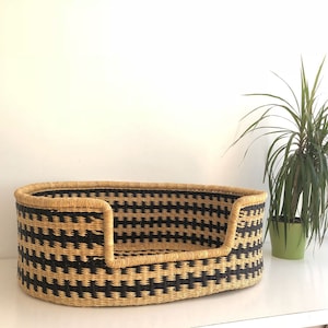 May include: A black and tan woven dog bed with a rounded shape. The bed is made of natural materials and has a raised rim.