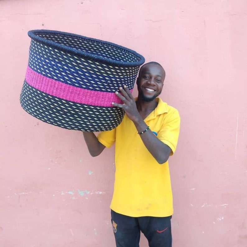 May include: A large, woven basket with a black, blue, and pink design. The basket has a cylindrical shape with a dark blue rim. The main body features a pattern of black and white woven material, with a pink band in the middle.