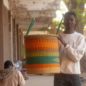 May include: A person holds a large, woven basket with orange, green, and brown stripes. The basket has two handles and is made of natural materials.