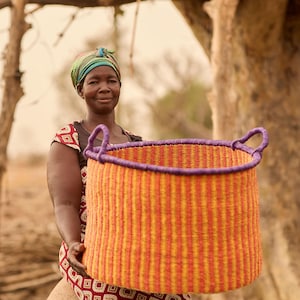 May include: A woman wearing a colourful headscarf holds a large, woven basket with orange and purple stripes. The basket has two handles and is made of natural materials.