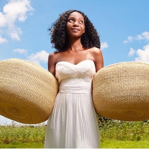 Puede incluir: Una mujer con un vestido blanco sostiene dos cestas grandes, redondas y tejidas frente a ella. Las cestas est&aacute;n hechas de materiales naturales y tienen un color marr&oacute;n claro. La mujer sonr&iacute;e y mira hacia el cielo. El fondo es un cielo azul con nubes blancas.