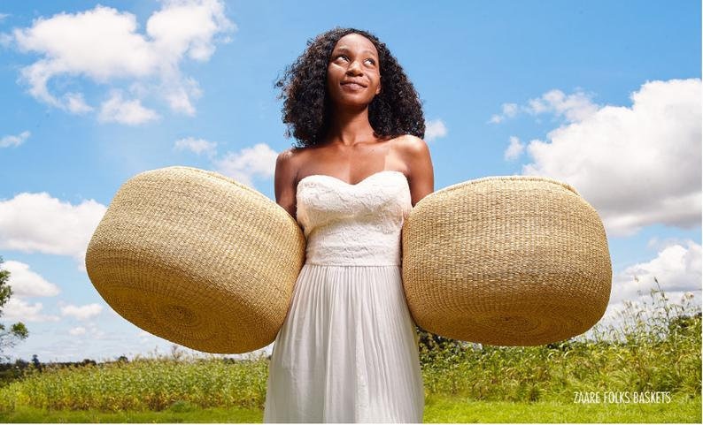 Puede incluir: Una mujer con un vestido blanco sostiene dos cestas grandes, redondas y tejidas frente a ella. Las cestas est&aacute;n hechas de materiales naturales y tienen un color marr&oacute;n claro. La mujer sonr&iacute;e y mira hacia el cielo. El fondo es un cielo azul con nubes blancas.