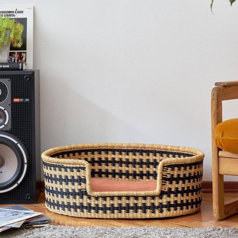 May include: A black and natural woven pet bed with a pink cushion. The bed is oval shaped and has a raised rim.