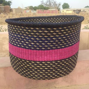 May include: A woven basket with a black rim and a curved top. The basket features a pattern of black, blue, and tan woven fibers, with a vibrant pink stripe. The basket is likely used for storage or decoration.