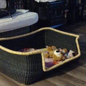 Puede incluir: Una cama para mascotas de mimbre negro con ribete beige. La cama está llena de una manta colorida y un animal de peluche marrón.