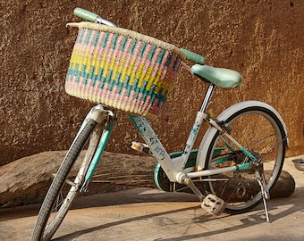 May include: A teal and white bicycle with a wicker basket. The basket is pink, yellow, and green. The bicycle has a kickstand and is parked on a dirt path.